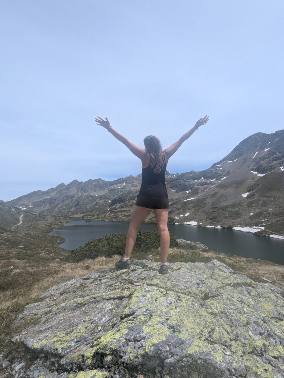 Person mit erhobenen Armen auf einem Felsen mit Blick auf eine Berglandschaft.
